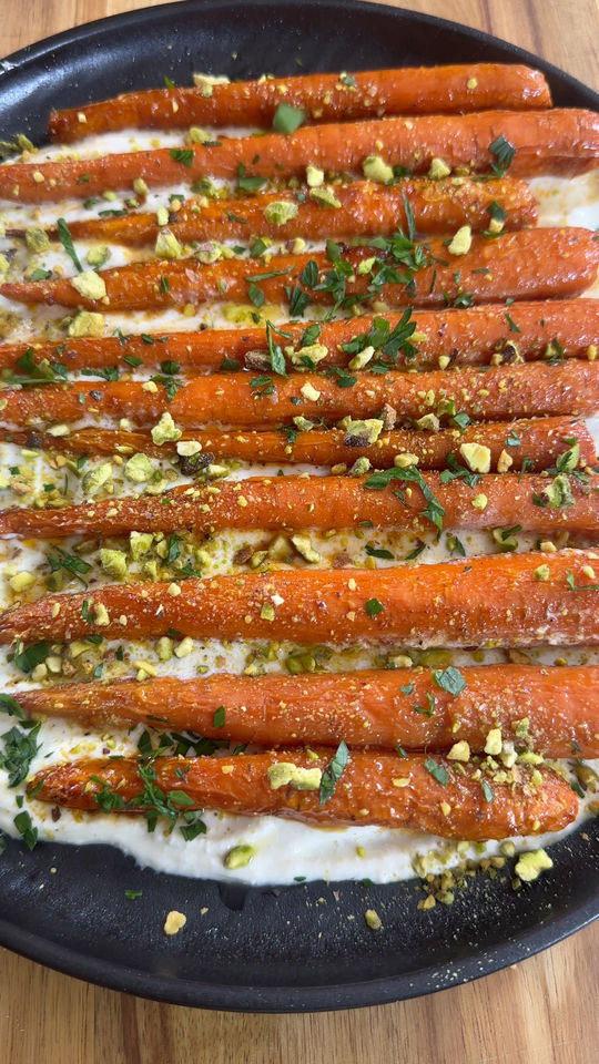 Roasted carrots with crushed pistachios and herbs on a bed of creamy sauce in a black dish.