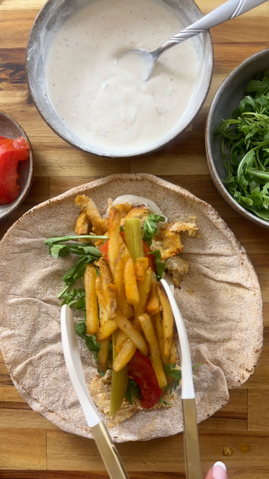 Preparing a pita wrap with fries, chicken, and greens on a wooden surface with sauce and salad bowls.
