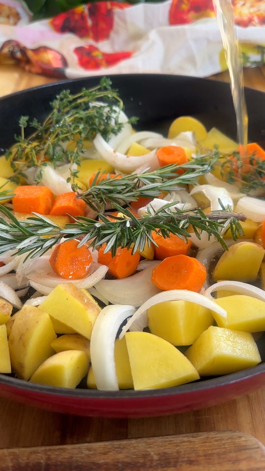 Pan with chopped potatoes, carrots, onions, rosemary, and thyme, being drizzled with broth.