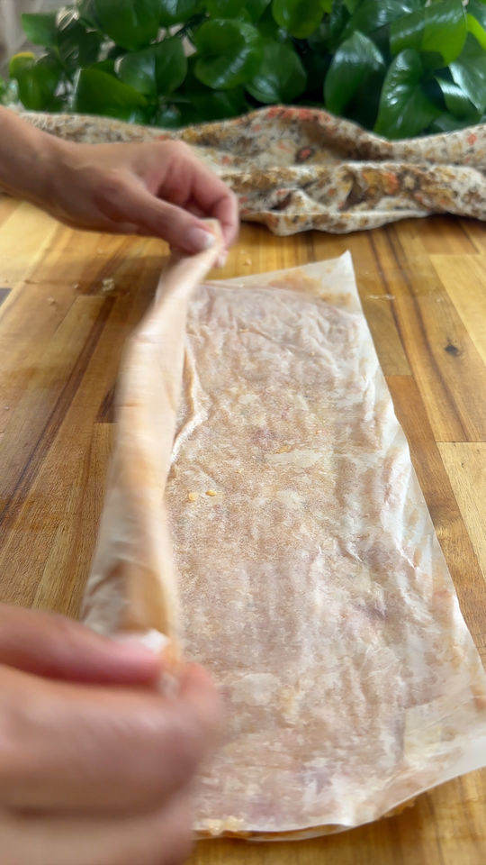 Hands rolling ground meat on a wooden surface with green leaves in the background.