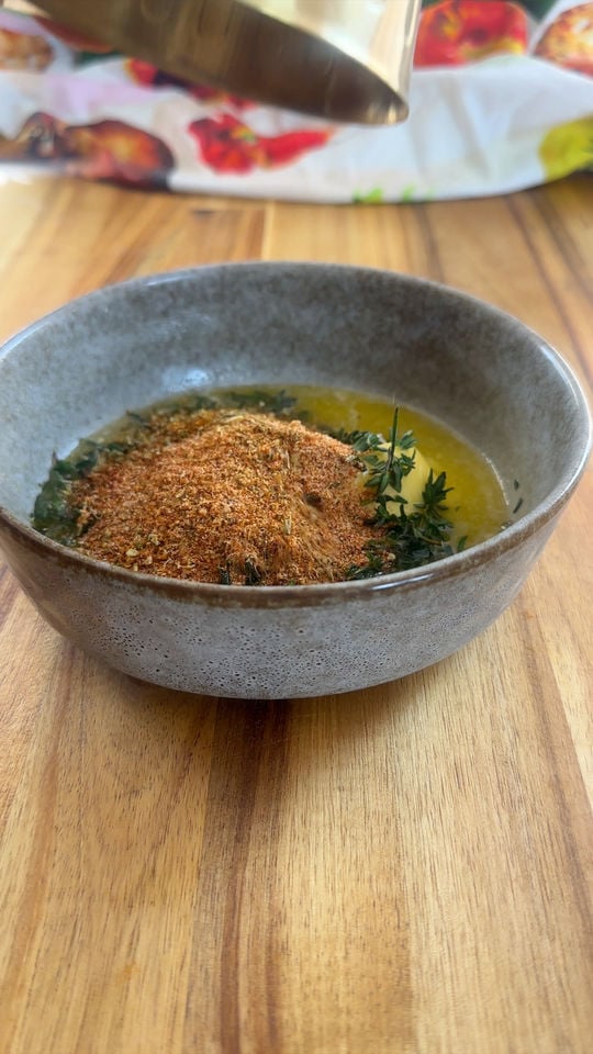 Bowl of spices, herbs, and oil on a wooden table, with a metal container above it.