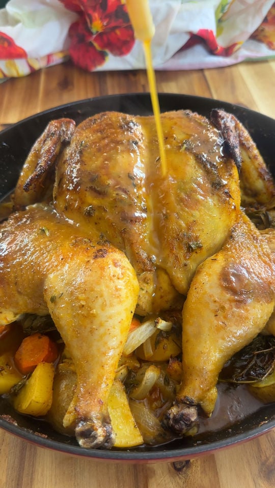 Roasted whole chicken with herb seasoning is being drizzled with sauce in a pan with vegetables.