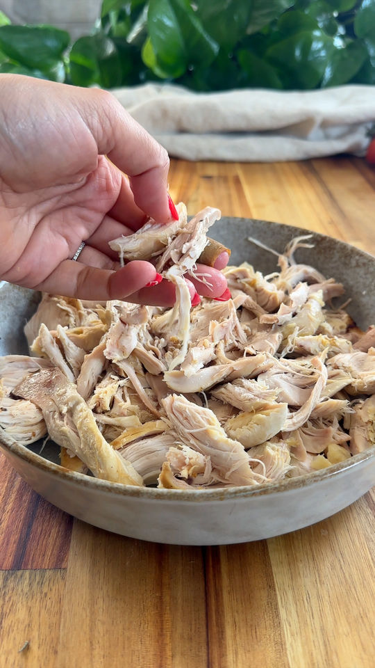 Hand shredding cooked chicken breast in a bowl on a wooden table.