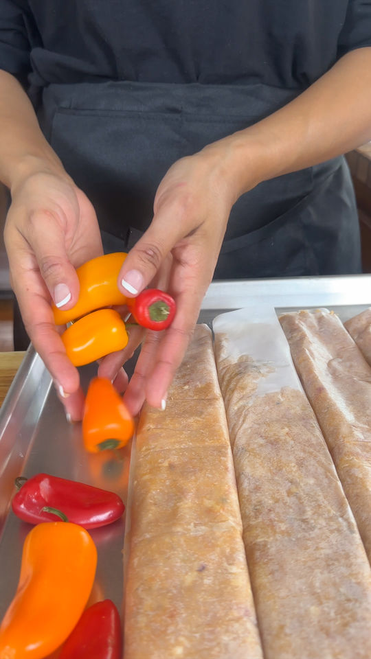 Hands arranging orange and red peppers next to wrapped meat logs on a metal tray.