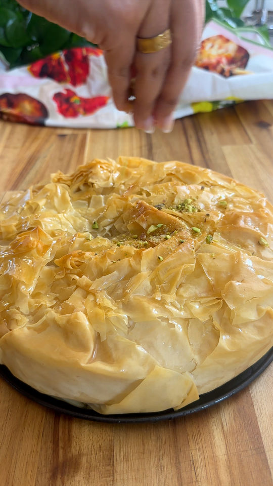 Hand sprinkling pistachios on a golden, flaky baklava pie on a black plate, wooden table underneath.