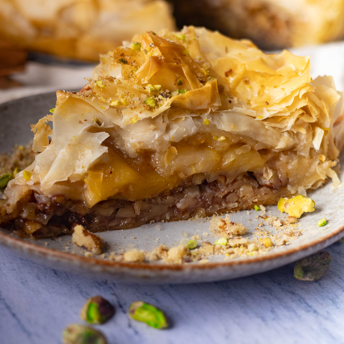 A slice of apple pie baklava with flaky phyllo layers, honey, and crushed pistachios on a plate.