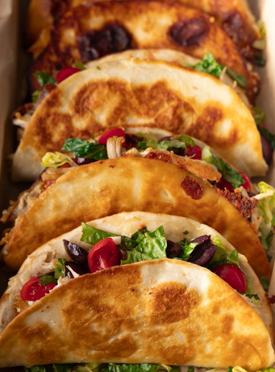 Golden brown Greek crispy chicken tacos filled with lettuce, tomatoes, and olives in a close-up view.
