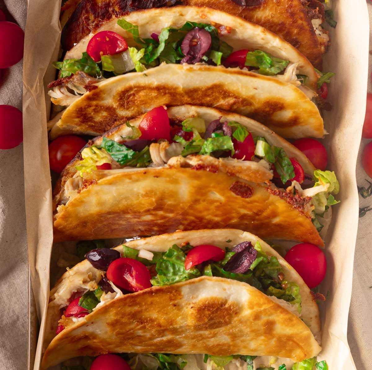 Greek crispy chicken tacos filled with lettuce, tomatoes, olives, and shredded chicken on a parchment-lined tray.