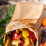 Viral doner kebab wrapped in pita in brown paper with grilled chicken, peppers, and fries on a plate with herbs.