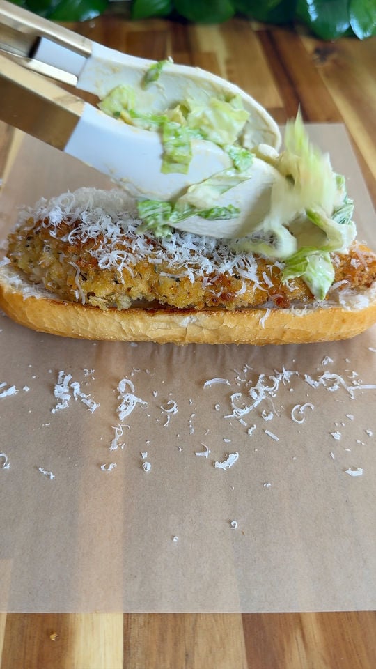 Tongs adding lettuce to a breaded cutlet sandwich topped with grated cheese on a wooden surface.