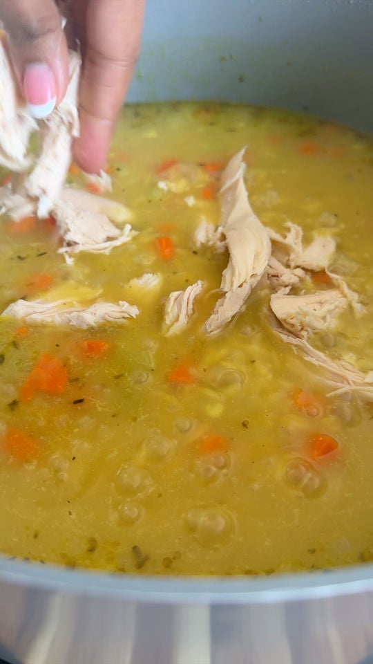 Hand adding shredded chicken to a pot of soup with carrots and herbs.