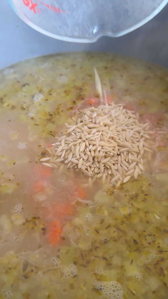 Pouring orzo into a pot of simmering soup with vegetables and herbs.