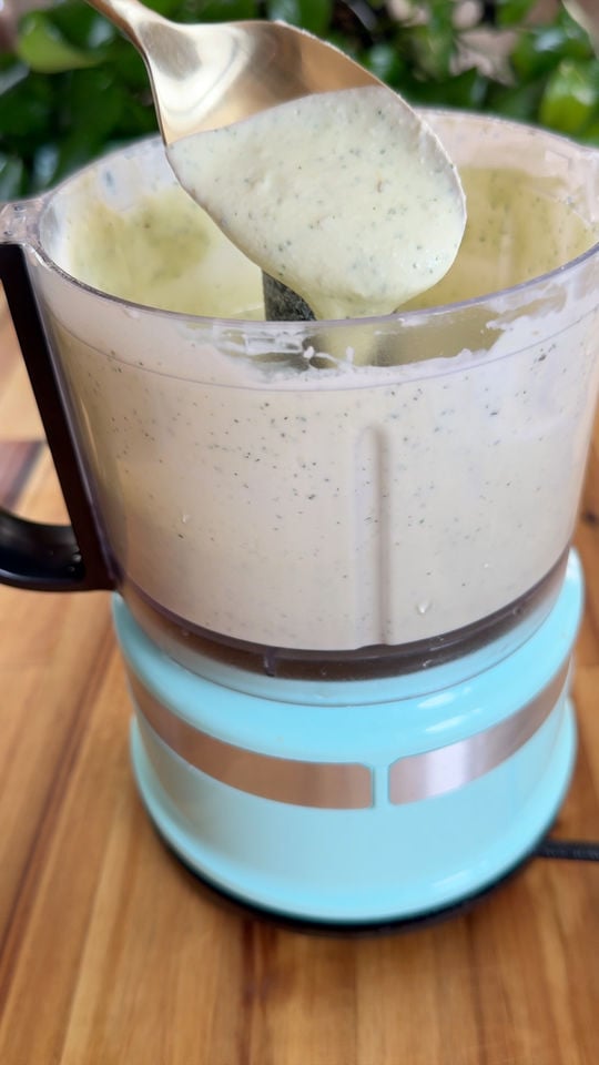 Food processor with creamy feta sauce being scooped by a spoon, set on a wooden countertop.