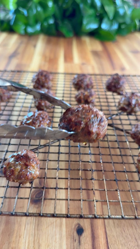 Tongs holding a meatball over a cooling rack with more meatballs on a wooden surface.
