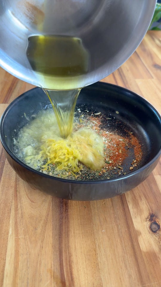 Pouring hot oil into a black bowl with spices and minced garlic on a wooden surface.