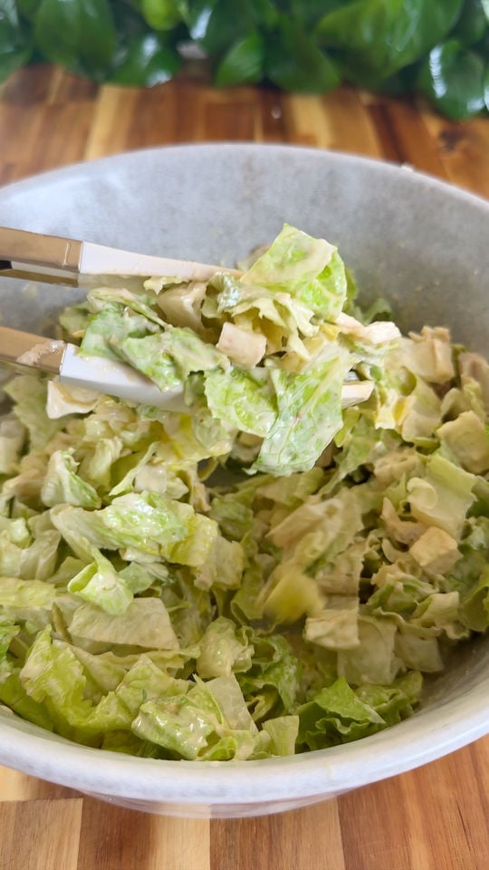 Tongs hold Caesar salad with lettuce and creamy dressing in a bowl on a wooden table.