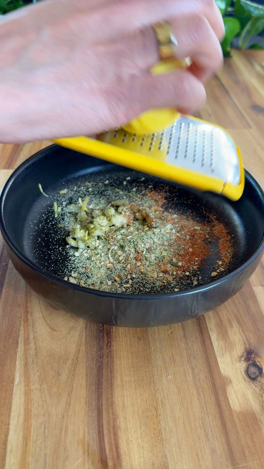 Hand grating lemon zest into a black bowl with spices on a wooden surface.