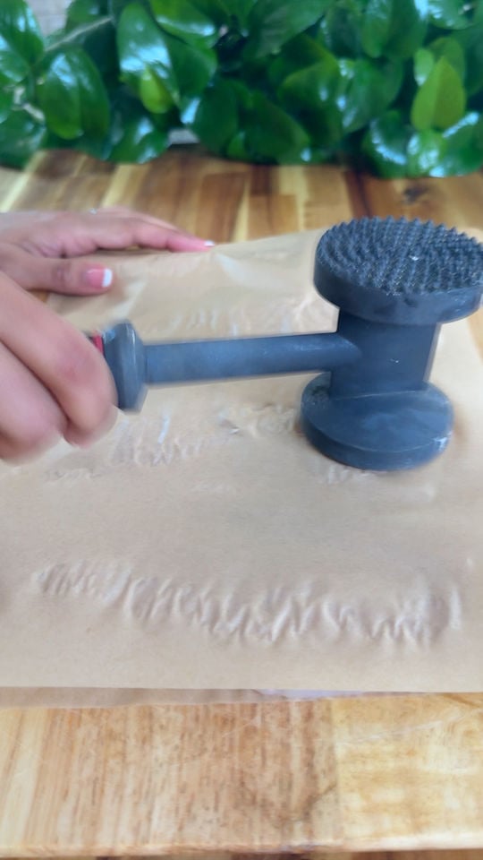 Hand using meat tenderizer on parchment paper over wooden surface.