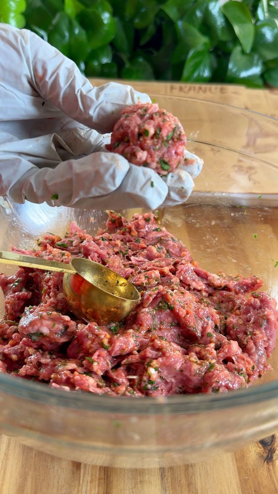 Gloved hands shaping seasoned ground lamb into a ball over a glass bowl on a wooden table.