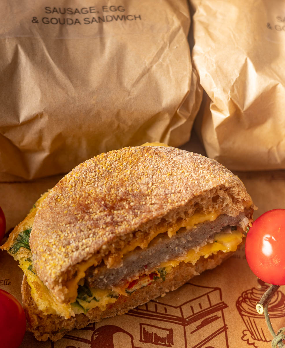 Sheet pan eggs breakfast sandwich with brown packaging in the background and tomatoes on the side.