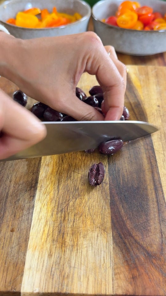 Hand slicing black olives on a wooden cutting board, with bowls of sliced peppers and yellow cherry tomatoes in the background.