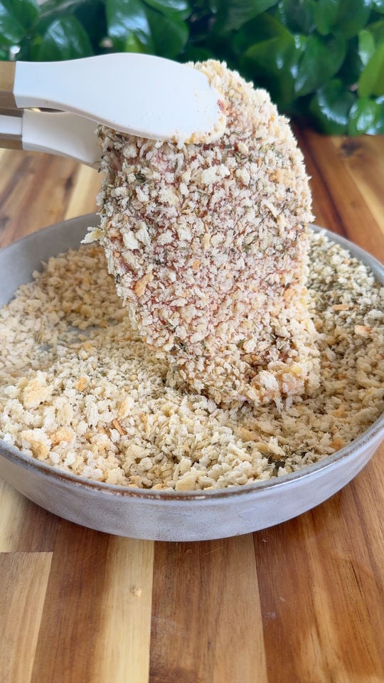 White tongs holding a bread crumb-coated chicken over a bowl of breadcrumbs on a wooden table.
