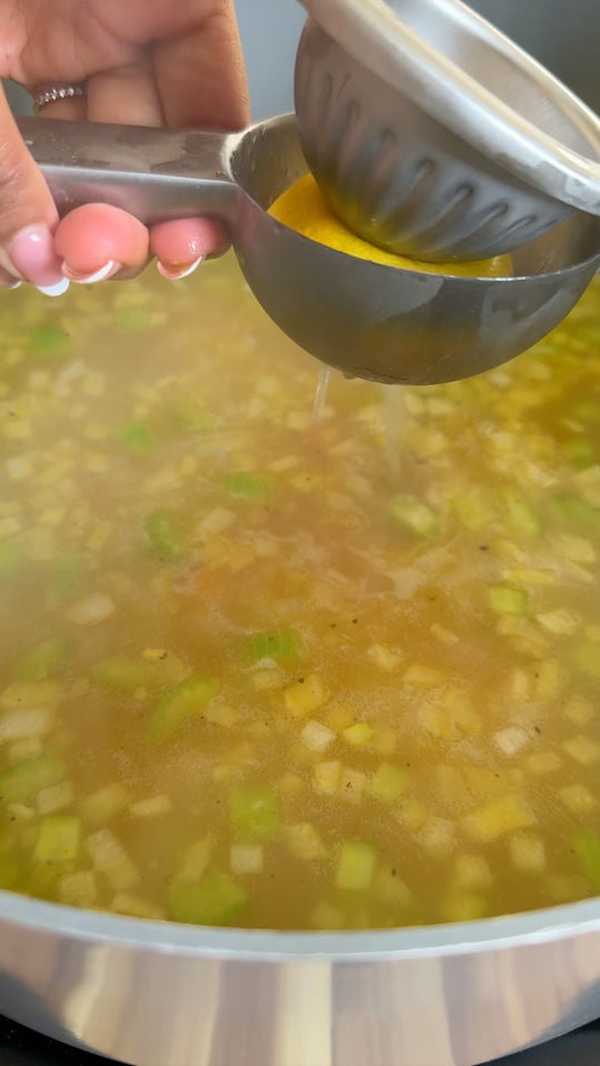 Hand squeezing juice from a lemon into a pot of simmering soup with chopped vegetables.