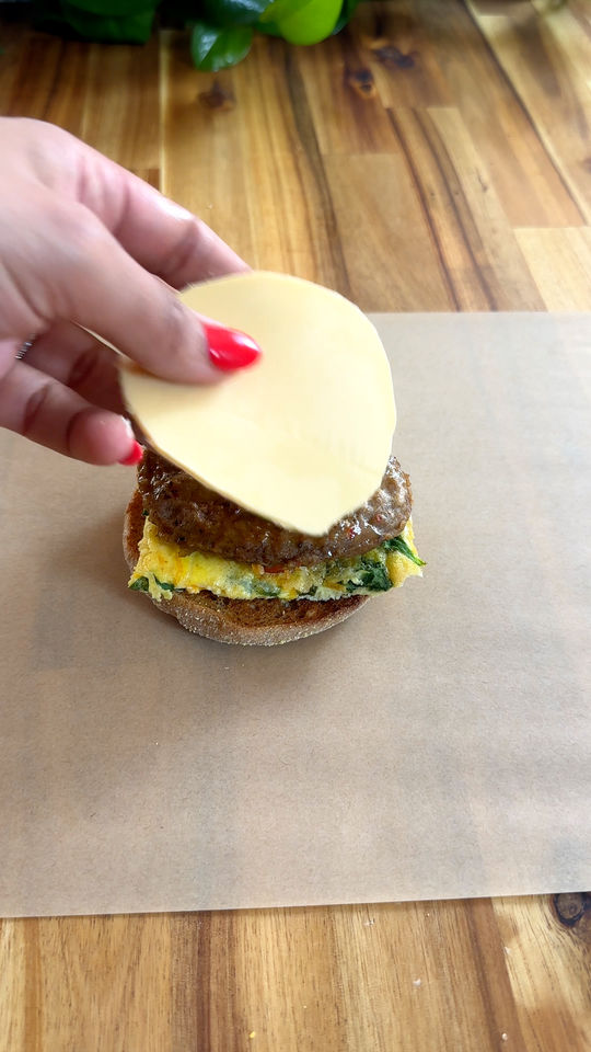 Hand placing cheese slice on a breakfast sandwich with egg and sausage on a wooden table.