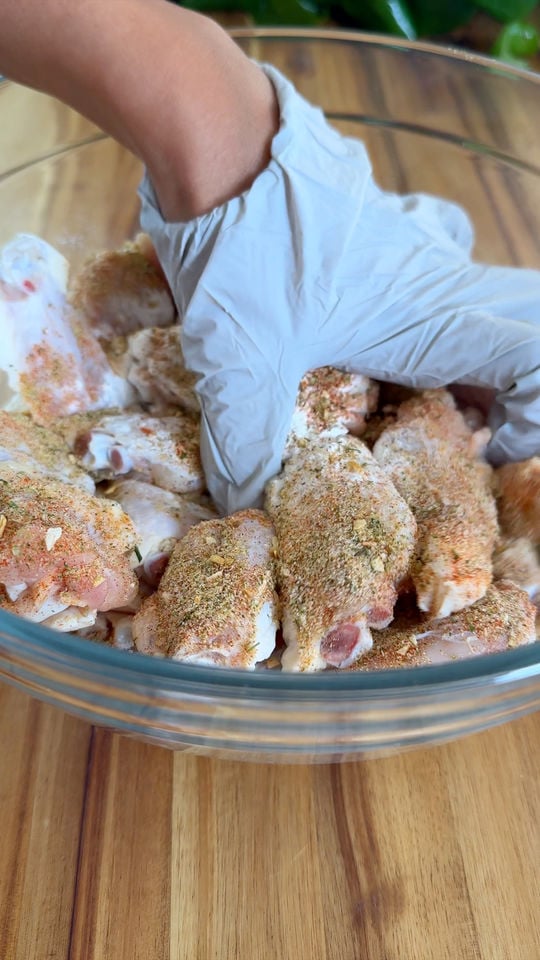 Hand in glove seasoning chicken wings in a glass bowl on a wooden surface.