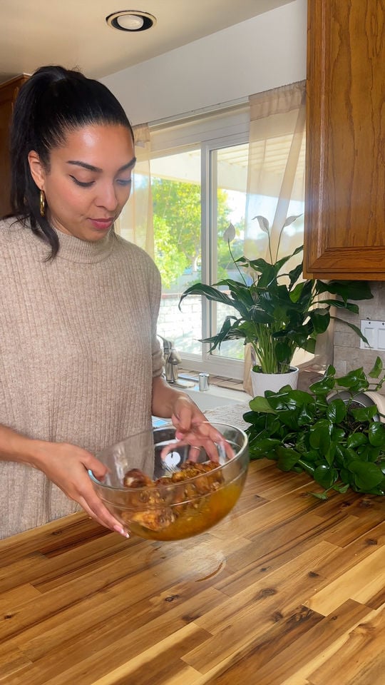Lola Jay in a kitchen holding a bowl of marinated chicken near a window with plants.