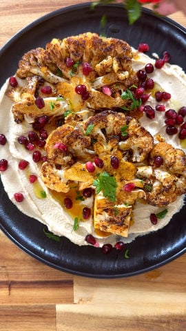 Roasted cauliflower on hummus, garnished with pomegranate seeds and herbs on a black plate.