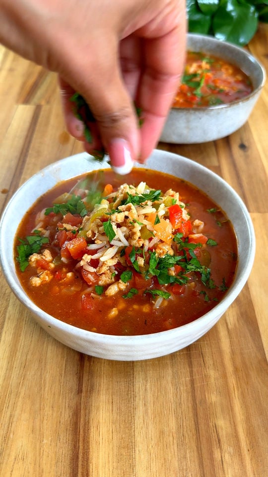 Hand garnishing soup with herbs in a white bowl on wooden table.