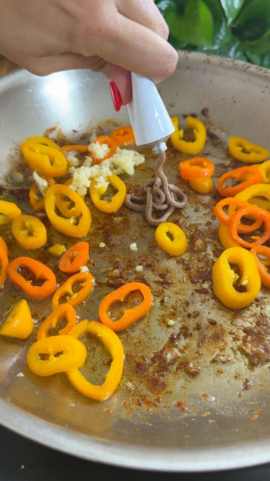 Hand adding paste into frying pan with yellow and orange bell pepper slices and chopped garlic.