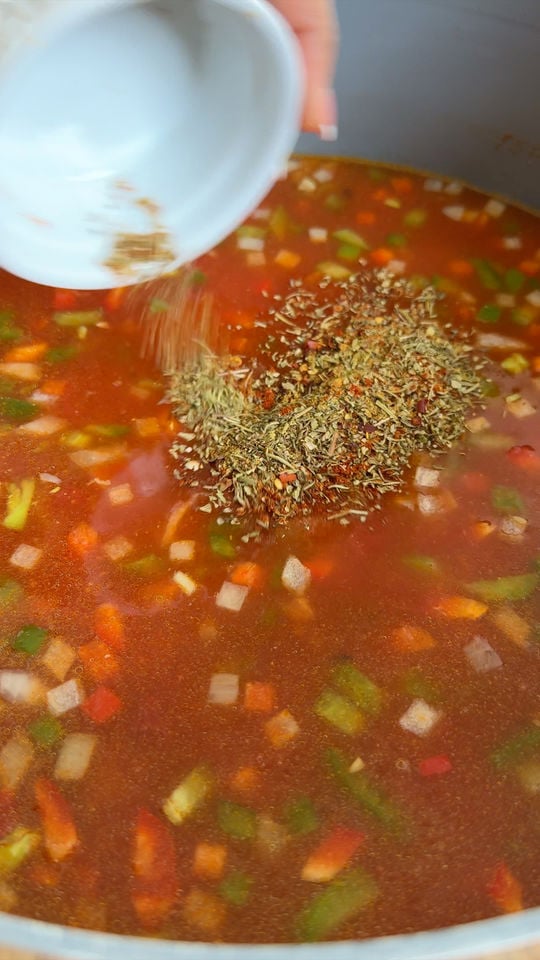 Adding spices to soup in a pot, with visible diced peppers and onions.