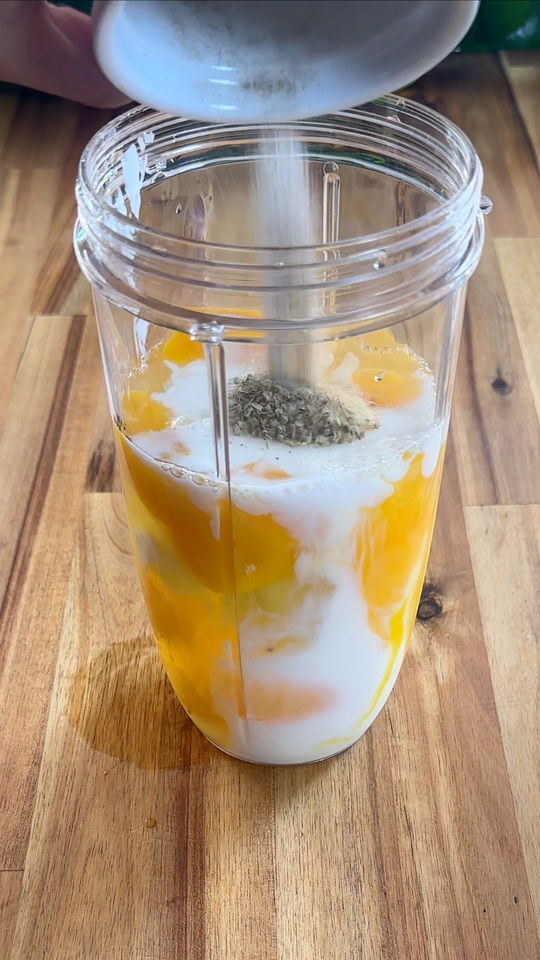 Eggs, milk, and spices being added to a blender cup on a wooden surface.