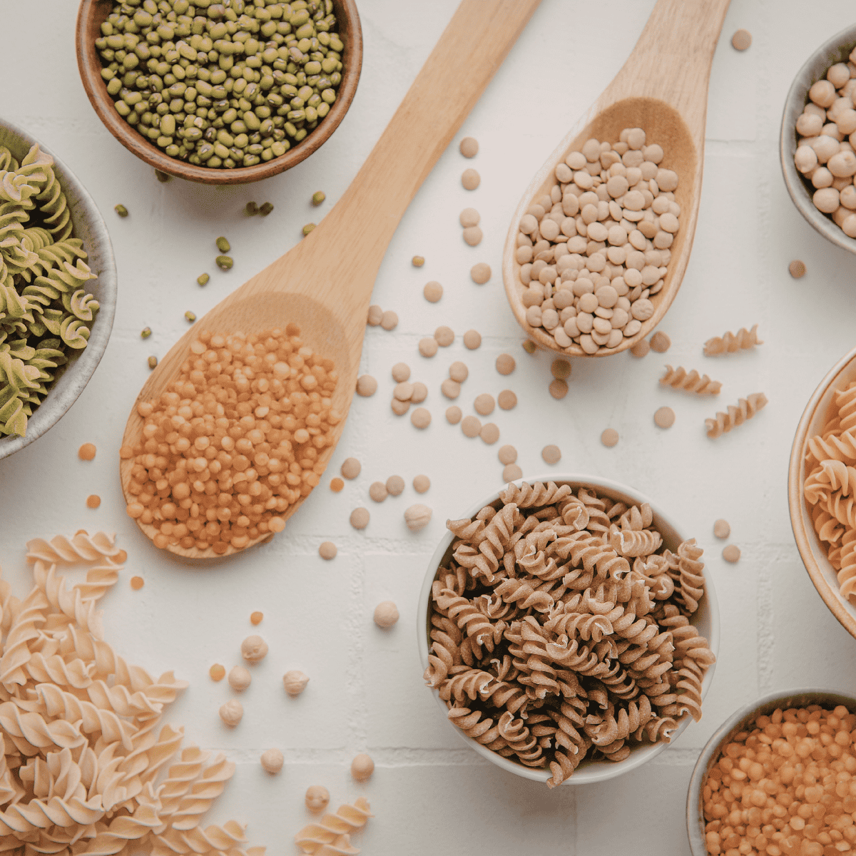 Assorted uncooked pasta types and legumes with wooden spoons on a white surface.