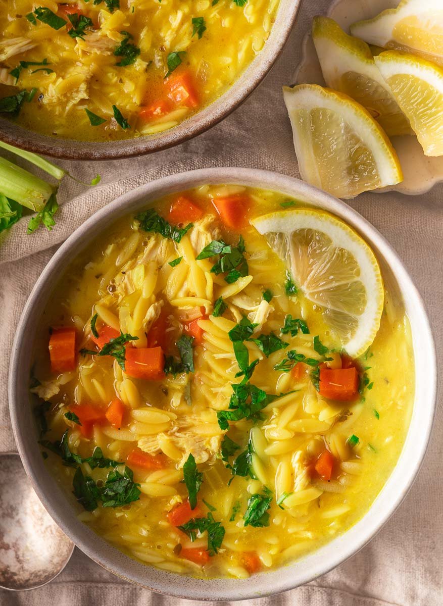 Bowl of chicken orzo soup with carrots, parsley, and lemon slices on a beige cloth, with a silver spoon beside.