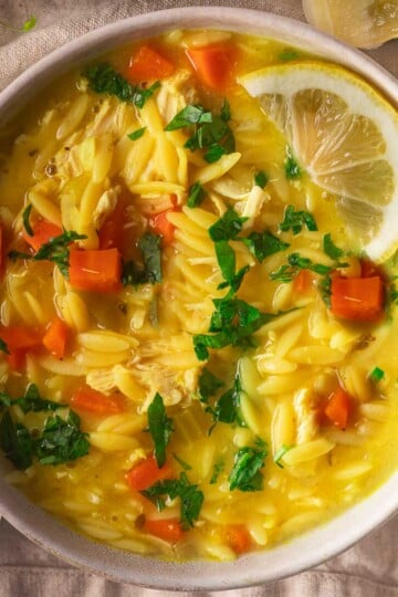 Lemon chicken orzo soup with carrots, lemon, and parsley garnish in a white bowl on a beige cloth.