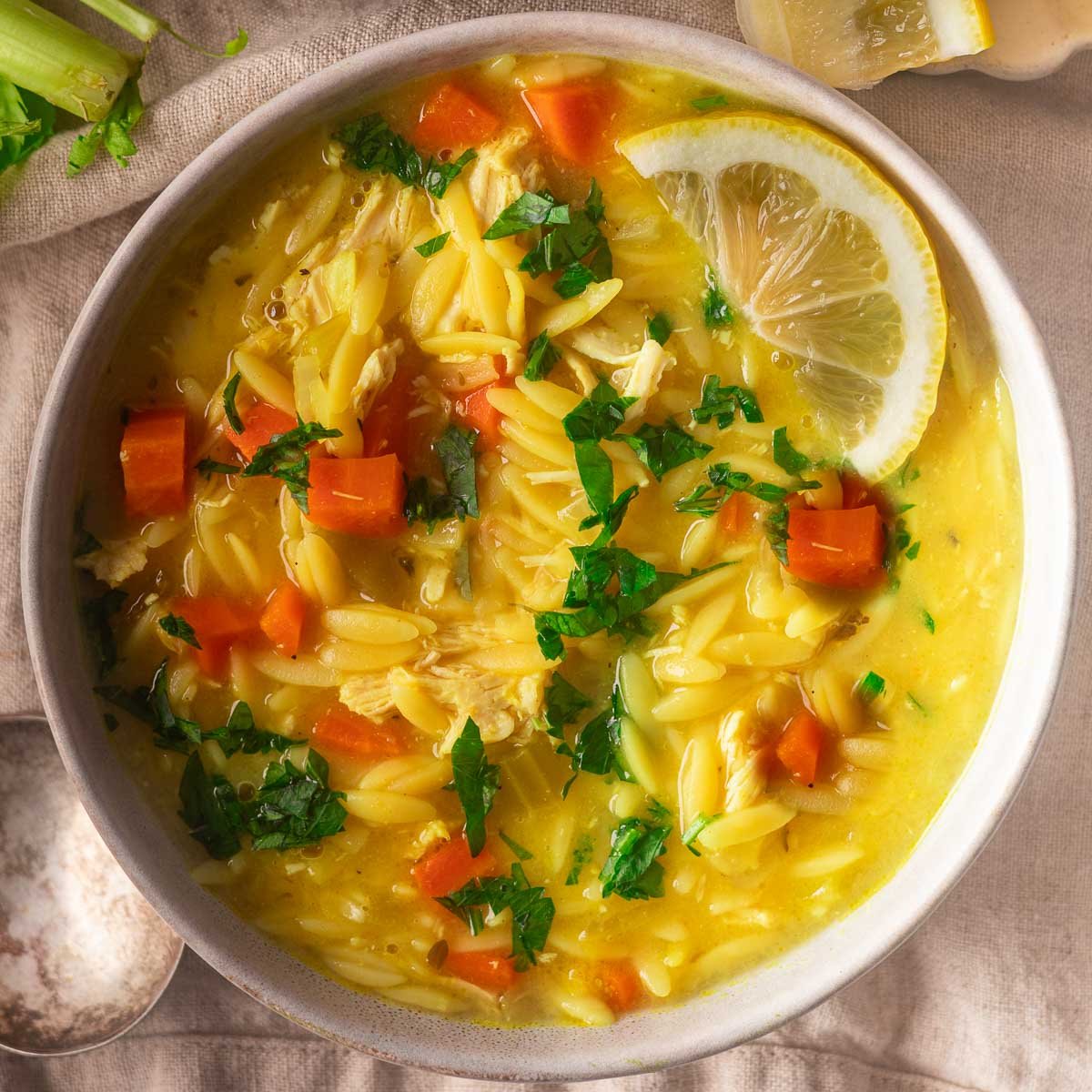 Lemon chicken orzo soup with carrots, lemon, and parsley garnish in a white bowl on a beige cloth.