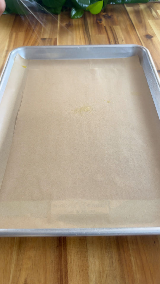 Baking sheet with parchment paper on wooden surface near green leaves.