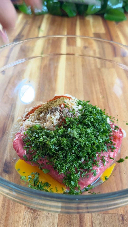 Ground lamb, chopped herbs, breadcrumbs, and spices in a glass bowl on a wooden surface.