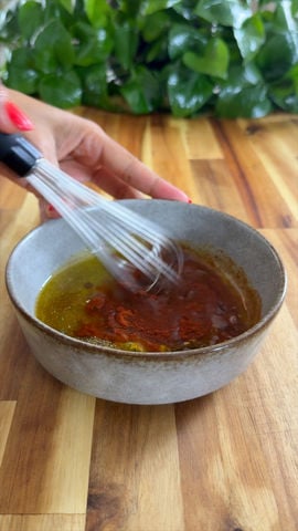 Whisk mixing spices and oil in a gray bowl on a wooden countertop with green leaves in the background.