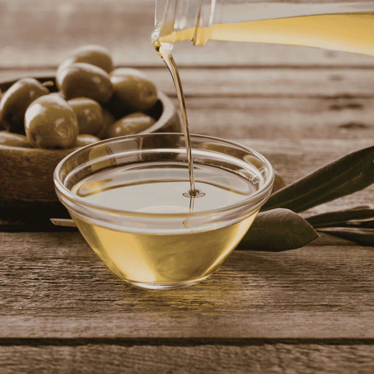Pouring olive oil into a glass bowl, with green olives in a wooden bowl on a rustic wooden table.
