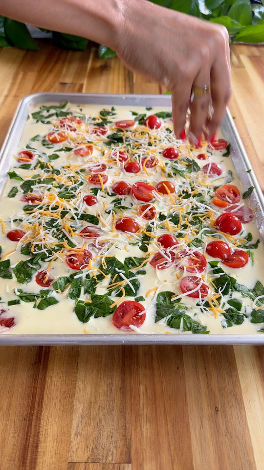 Hand placing cherry tomatoes and cheese on egg and spinach mixture in a baking tray on a wooden table.
