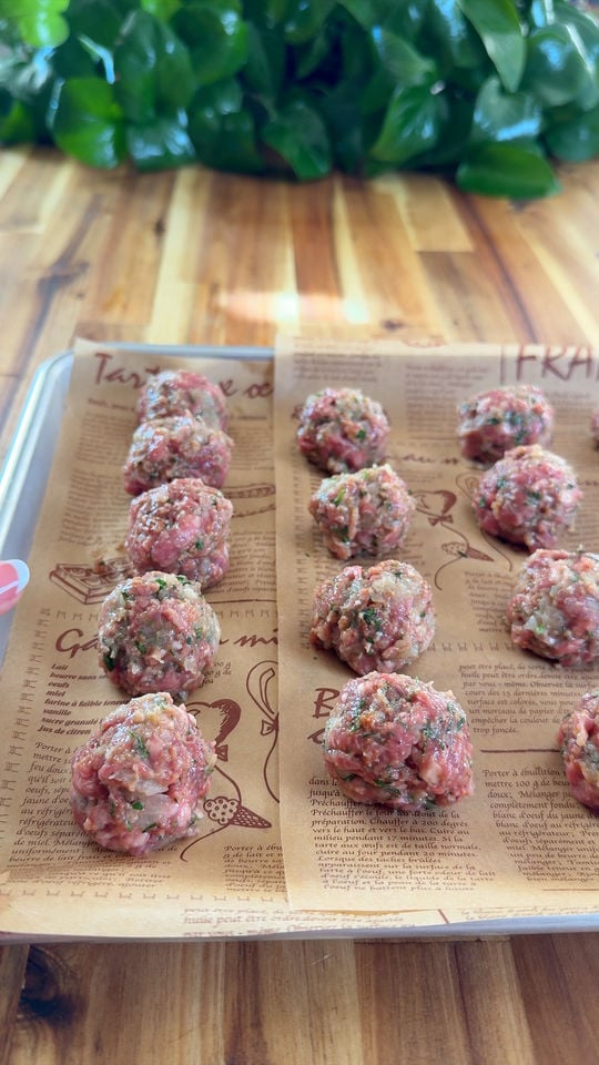 Raw meatballs arranged on parchment-lined tray, wooden surface, green leaves in background.