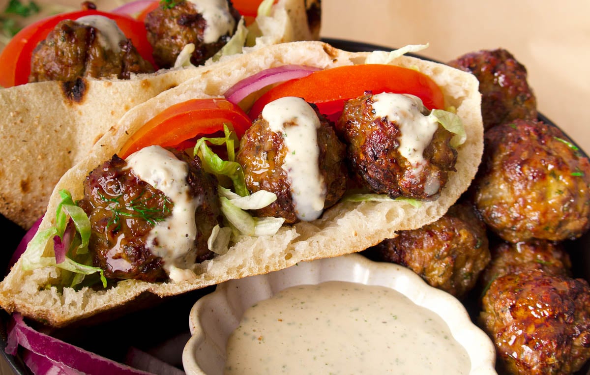 Spiced lamb meatballs with lettuce, tomato, red onion, and sauce, next to a bowl of creamy dipping sauce.
