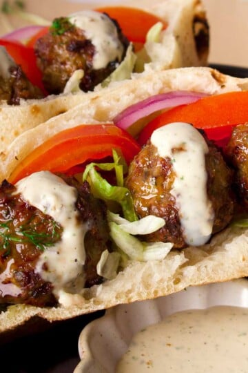 Pita sandwich with meatballs, lettuce, tomato, onion, drizzled with sauce, with a small dish of extra sauce.