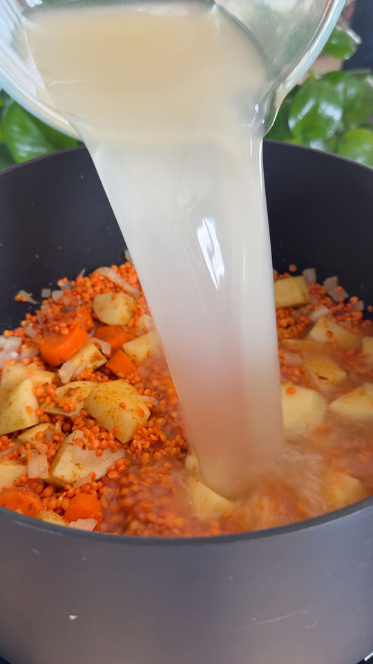Pouring broth into pot with potatoes, carrots, onions, and red lentils.