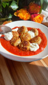 Meatballs with ricotta in red sauce on a white plate, garnished with bright flowers in the background.