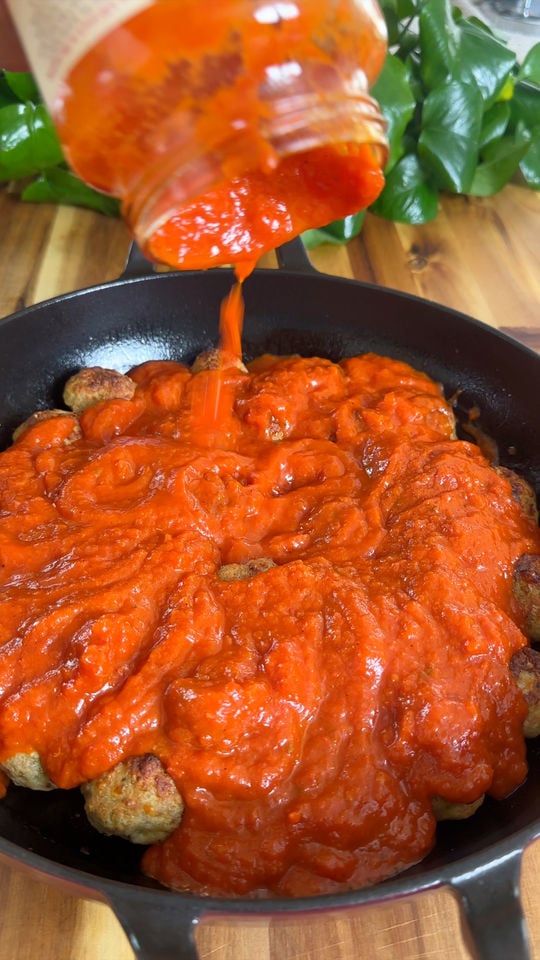 Pouring tomato sauce over meatballs in a black skillet with a green plant in the background.